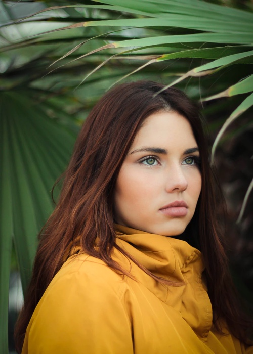 A beautiful young woman, with blue eyes and reddish brown hair wearing a yellow jacket surrounded by greenery
