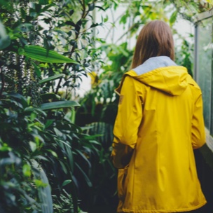 A brown haired person wearing a yellow rainjacket with the camera facing her back, staring into the greenery surrounding her